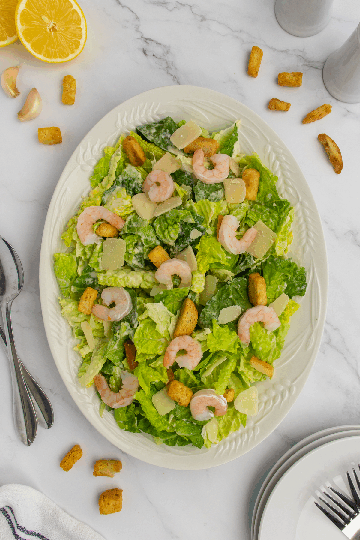 A large white plate with romaine lettuce, shrimp, croutons, and shaved parmesan cheese, set on a marble surface with lemon halves, garlic cloves, utensils, and plates nearby.