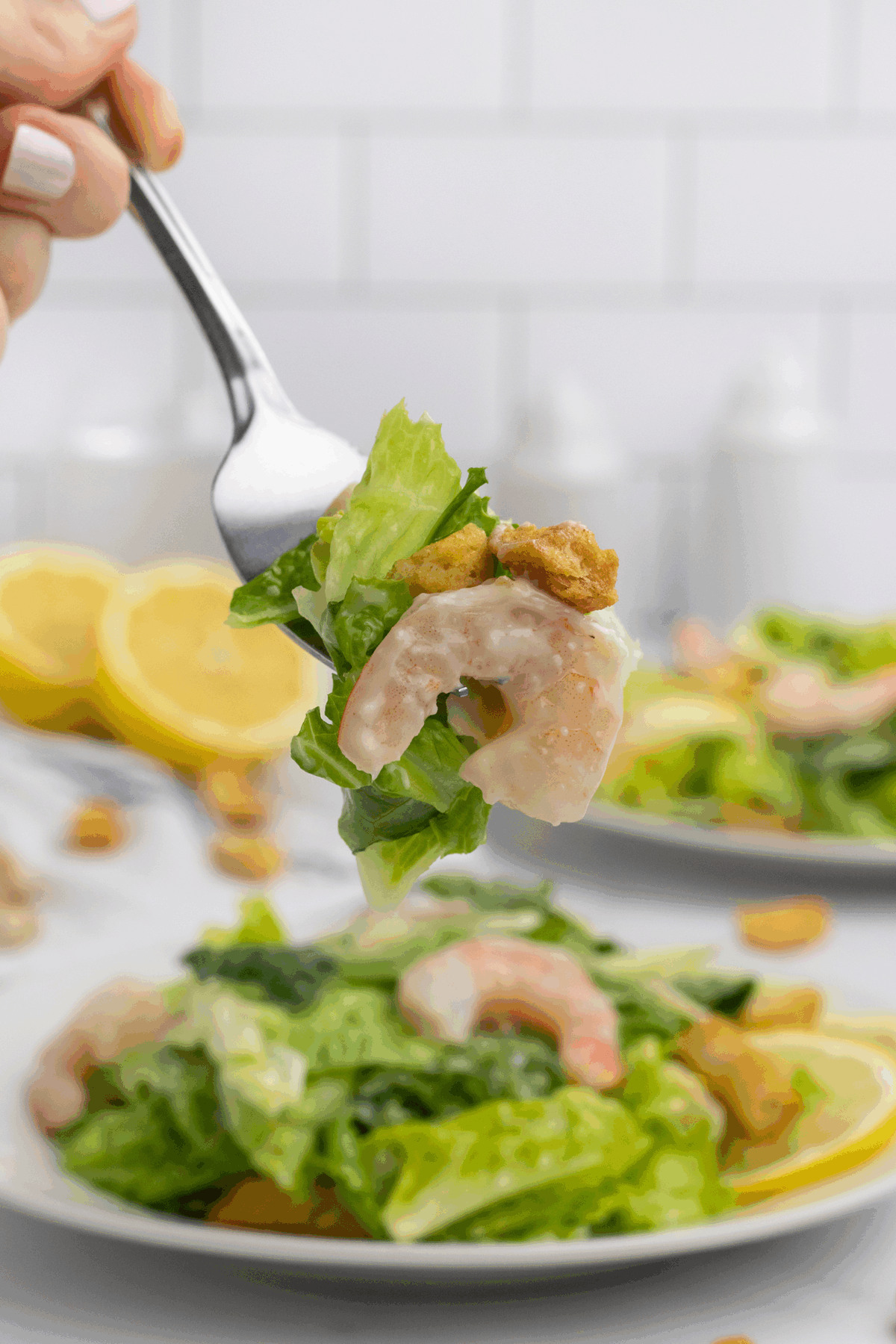 A fork holds up a bite of Caesar salad with shrimp, lettuce, croutons, and creamy dressing. In the background, a plate of similar salad and lemon halves are visible on a white surface.