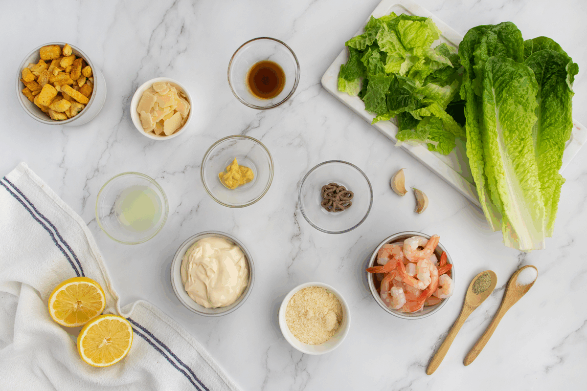 Overhead view of Caesar salad ingredients on a white surface: romaine lettuce, shrimp, mayonnaise, lemon halves, croutons, Parmesan, mustard, lemon juice, anchovies, garlic, spices, and sauce in small bowls.
