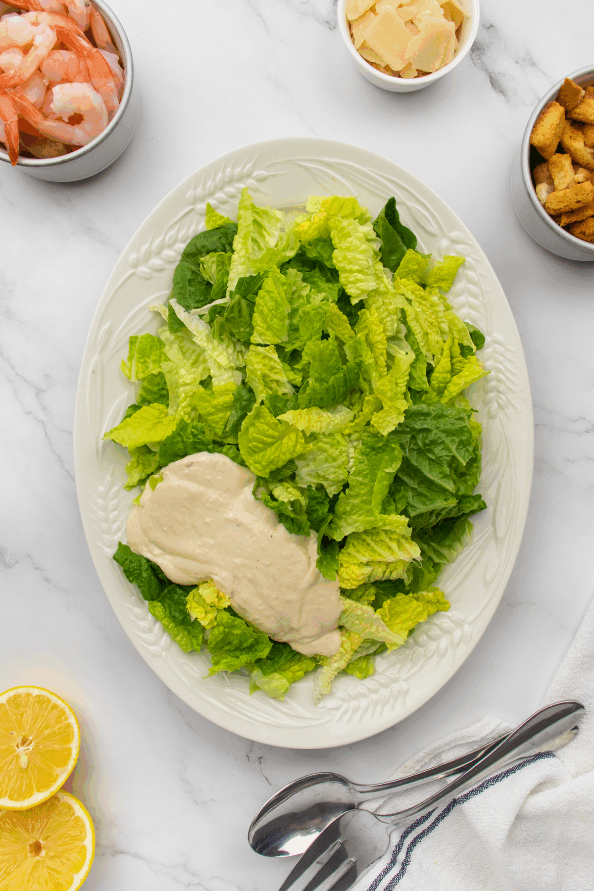 A plate of chopped romaine lettuce with Caesar dressing, surrounded by bowls of shrimp, Parmesan cheese, croutons, lemon halves, and utensils on a white marble surface.