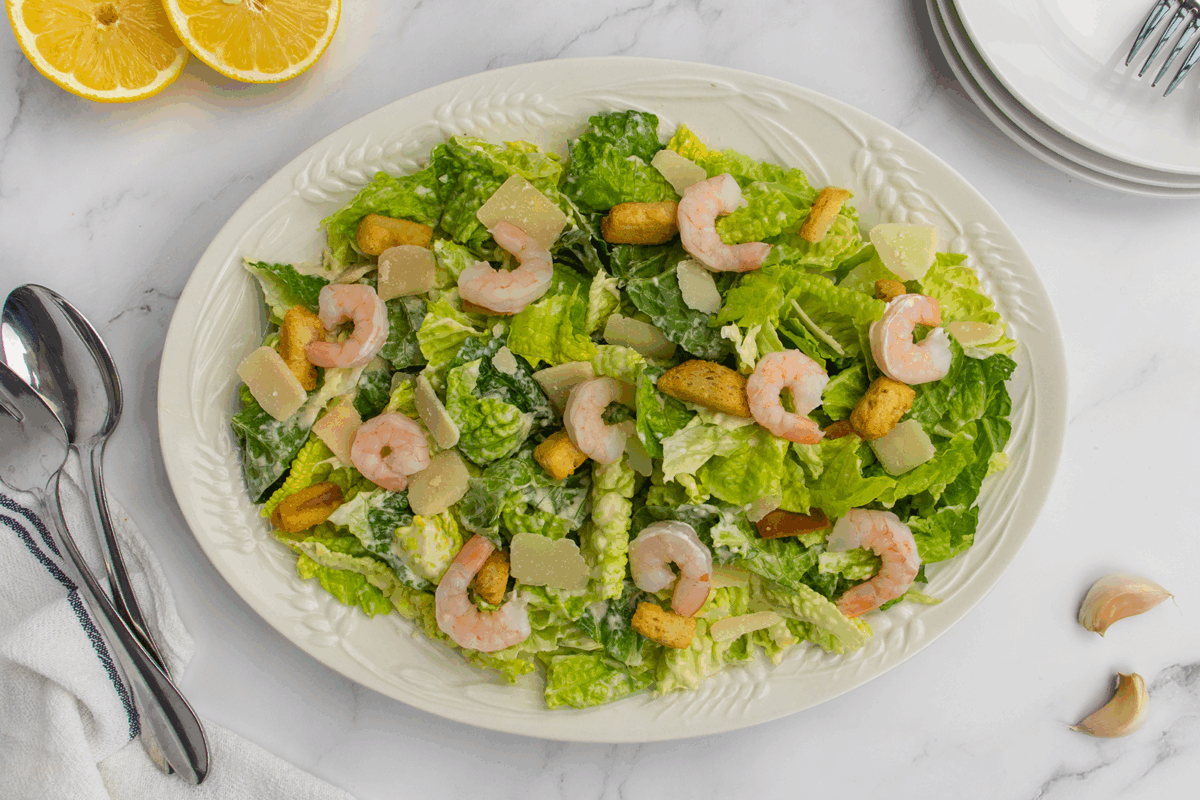 A white oval platter holds a Caesar salad topped with shrimp, croutons, and parmesan cheese on a marble surface. Lemon halves, silverware, plates, and garlic cloves are nearby.