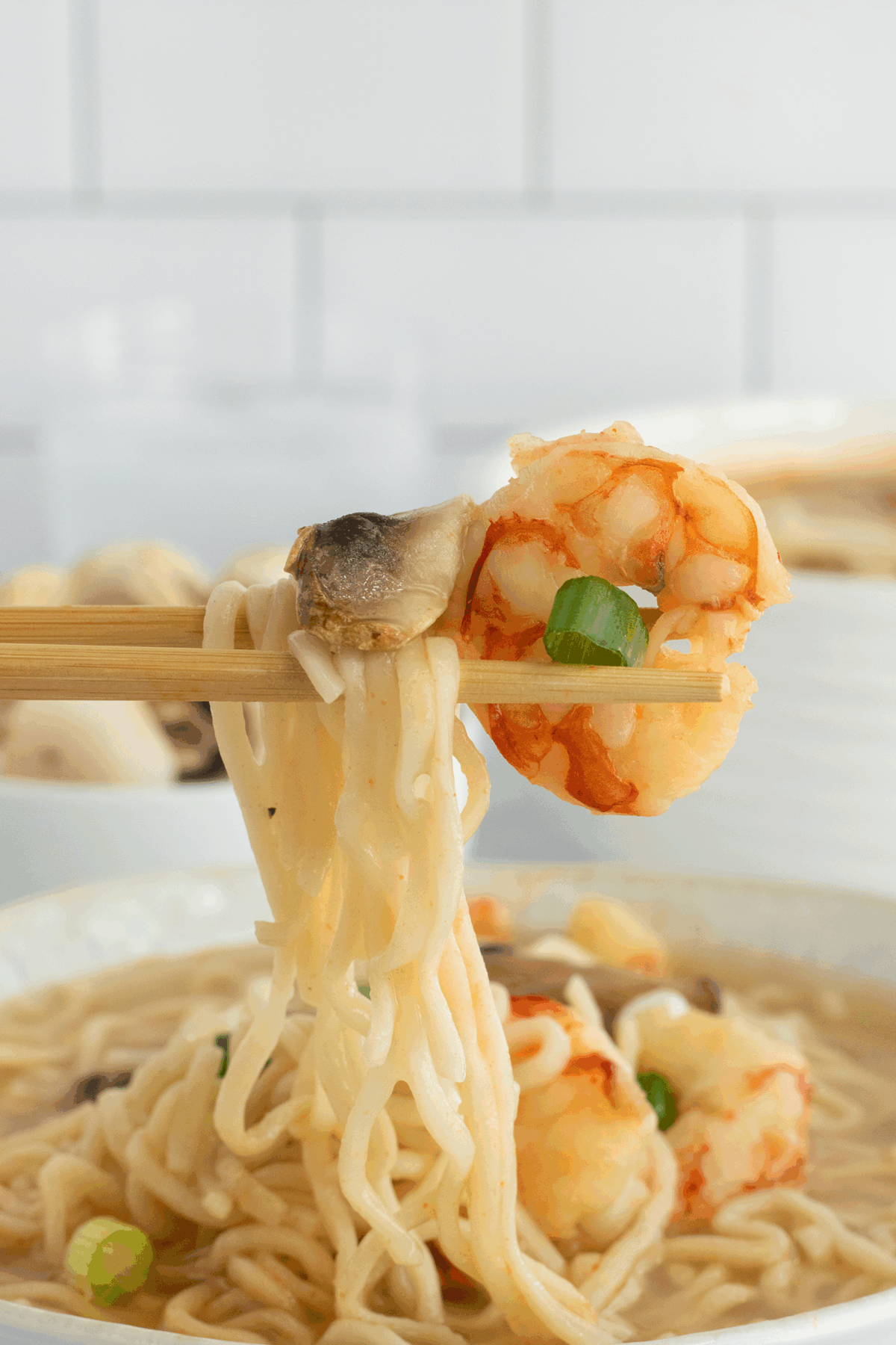 A pair of chopsticks holding up ramen noodles, a shrimp, a slice of mushroom, and a green onion above a bowl of soup.