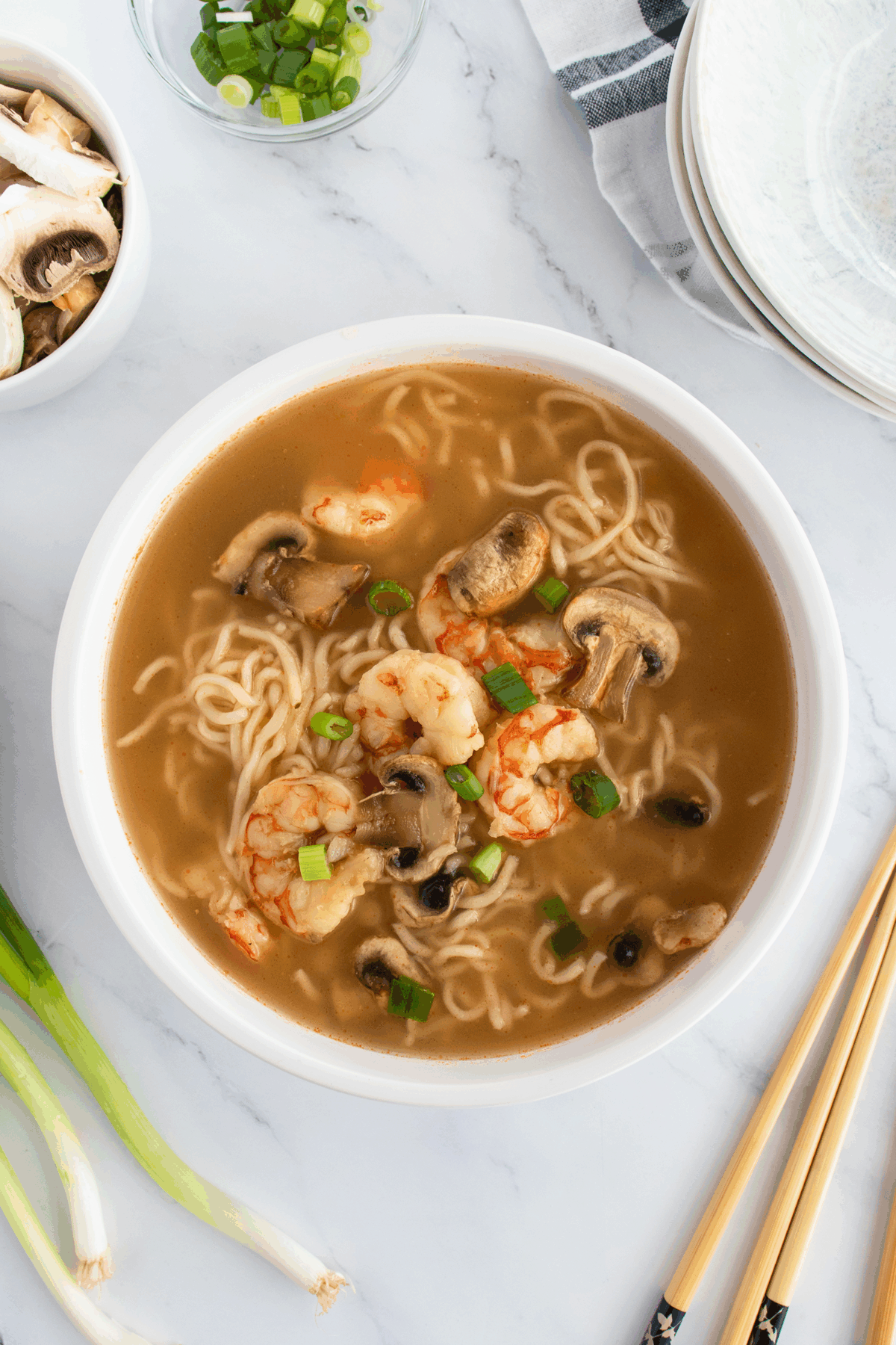 A bowl of ramen soup with shrimp, sliced mushrooms, and chopped green onions on a marble surface, with chopsticks, green onions, sliced mushrooms, and stacked plates nearby.