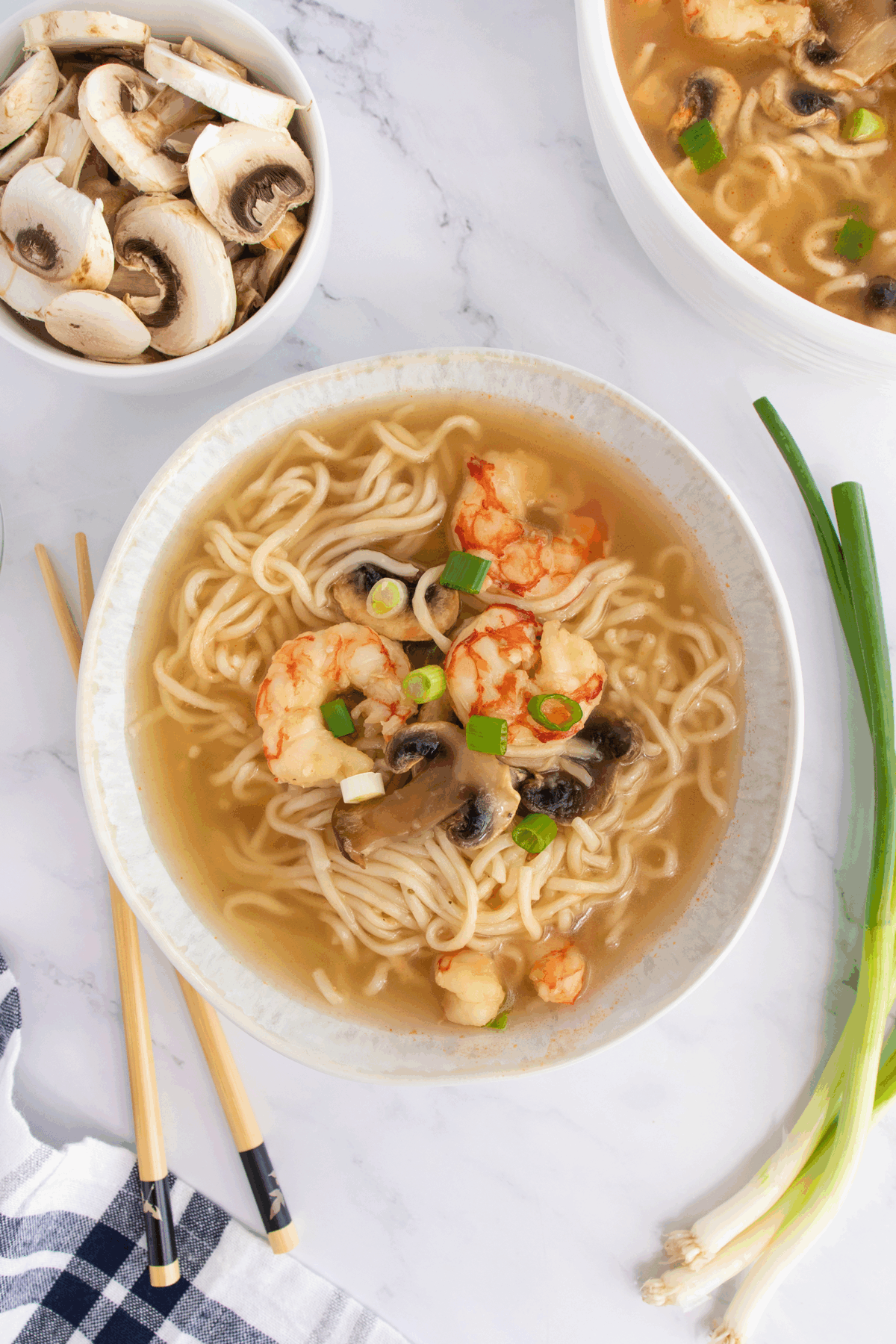 A bowl of noodle soup with shrimp, sliced mushrooms, and chopped green onions sits on a white surface. Nearby are raw mushroom slices, green onions, chopsticks, and another bowl of soup.