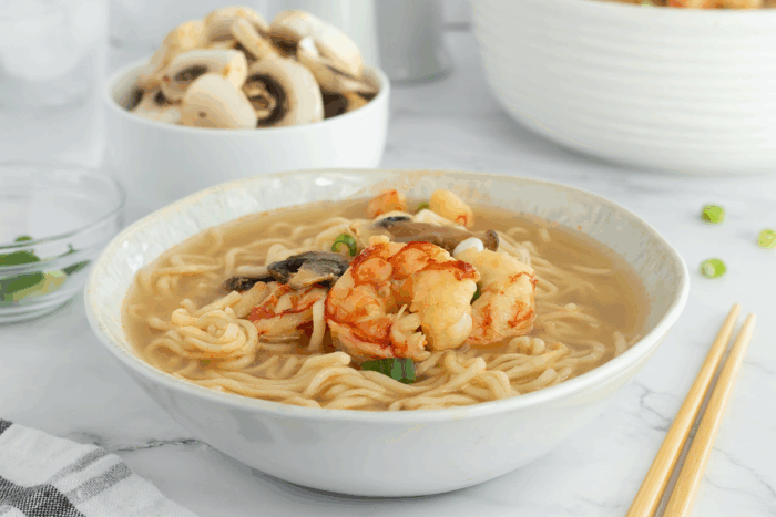 A bowl of noodle soup with shrimp and mushrooms, garnished with green onions. Sliced mushrooms and chopped green onions are in small bowls in the background, with chopsticks and a napkin beside the bowl.
