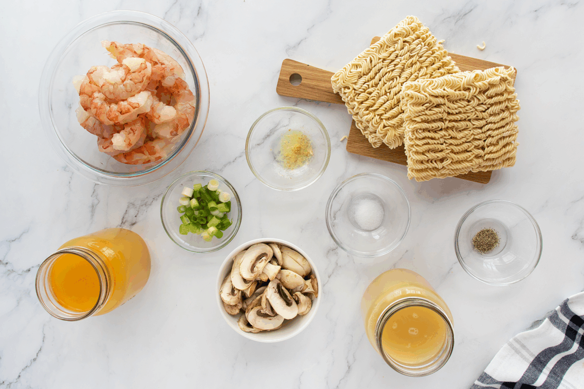 A top view of ingredients for shrimp ramen on a marble surface: raw shrimp in a bowl, dried ramen noodles, sliced mushrooms, chopped green onions, minced garlic, chicken broth, and seasonings in small bowls.