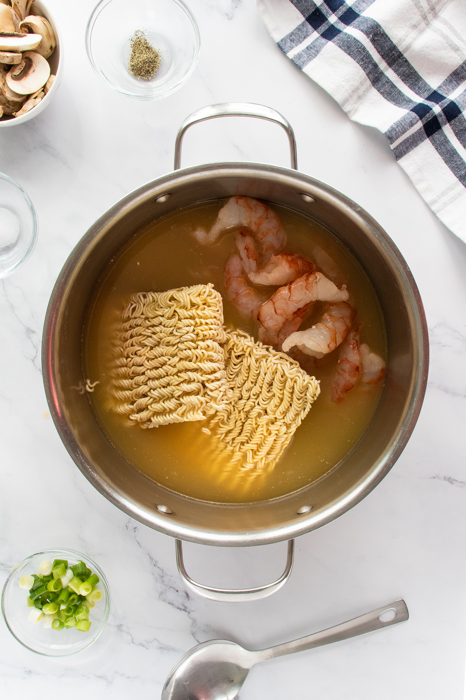 A pot of broth with uncooked instant ramen noodles and raw shrimp, surrounded by ingredients like sliced mushrooms, green onions, and seasonings on a white countertop.