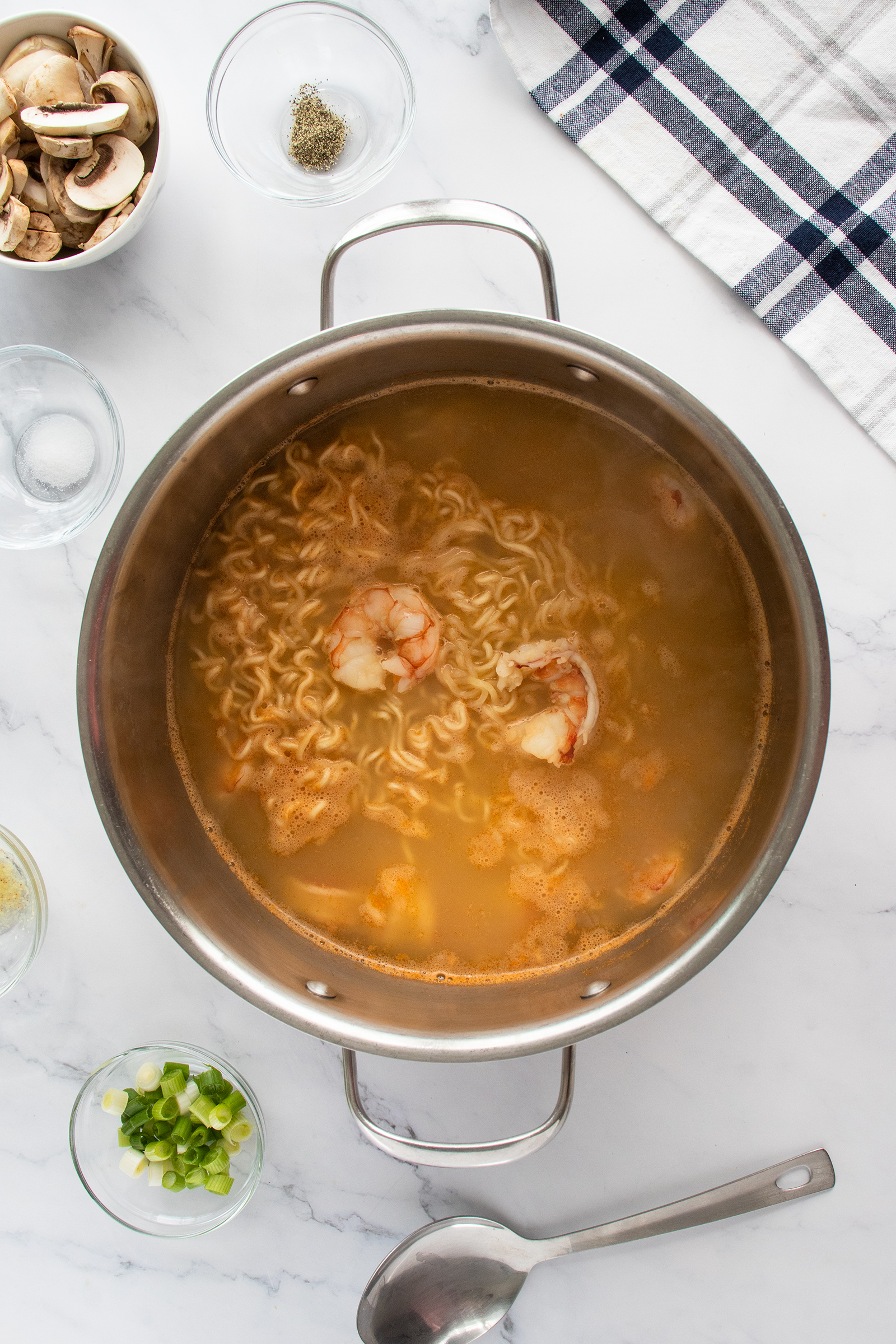 A pot of shrimp ramen soup with noodles and two shrimp in broth, surrounded by small bowls of green onions, mushrooms, pepper, and a metal ladle on a white marble surface with a checkered towel.