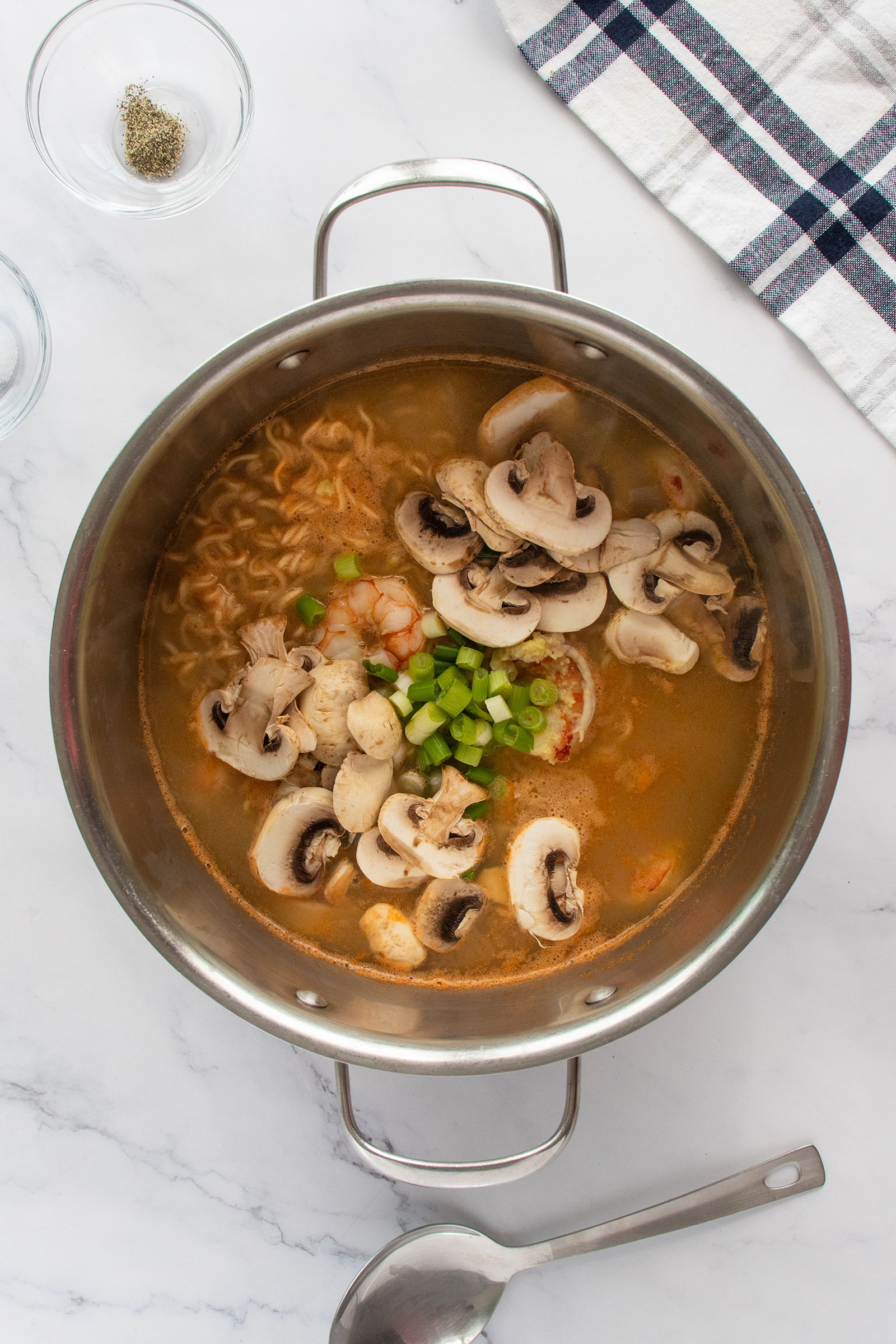A stainless steel pot filled with soup, containing ramen noodles, shrimp, sliced mushrooms, and chopped green onions, sits on a marble countertop next to a spoon and a white towel with dark stripes.