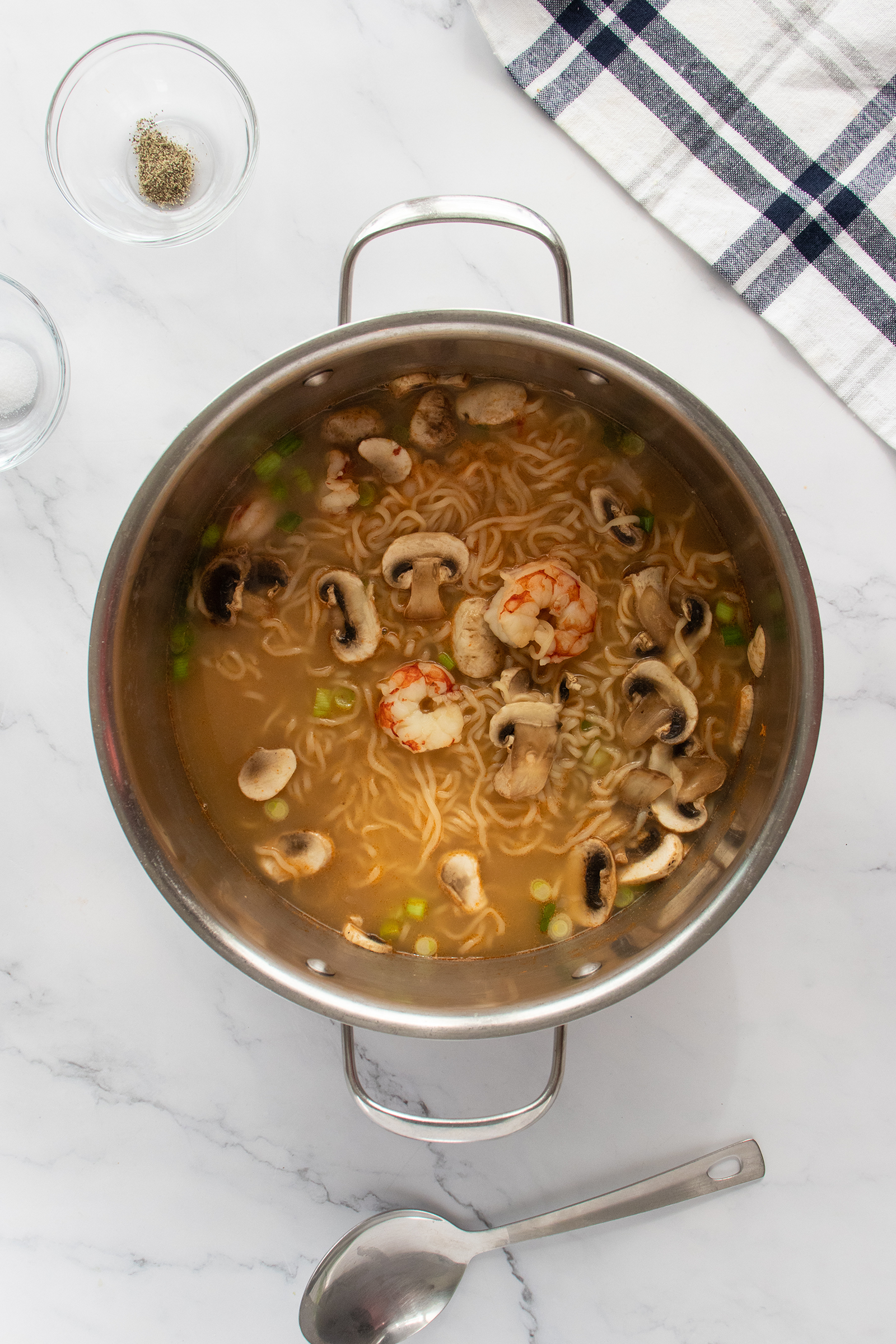 A pot of noodle soup with shrimp, sliced mushrooms, and green peas on a marble countertop. A spoon, glass bowls, and a folded towel are nearby.