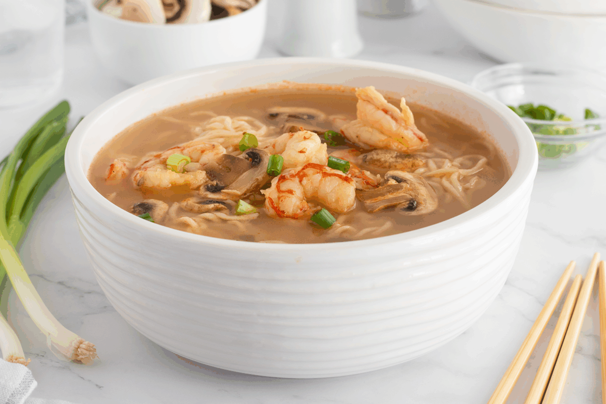A white bowl filled with shrimp and mushroom ramen soup, garnished with chopped green onions, sits on a marble surface with chopsticks, scallions, and dishes in the background.