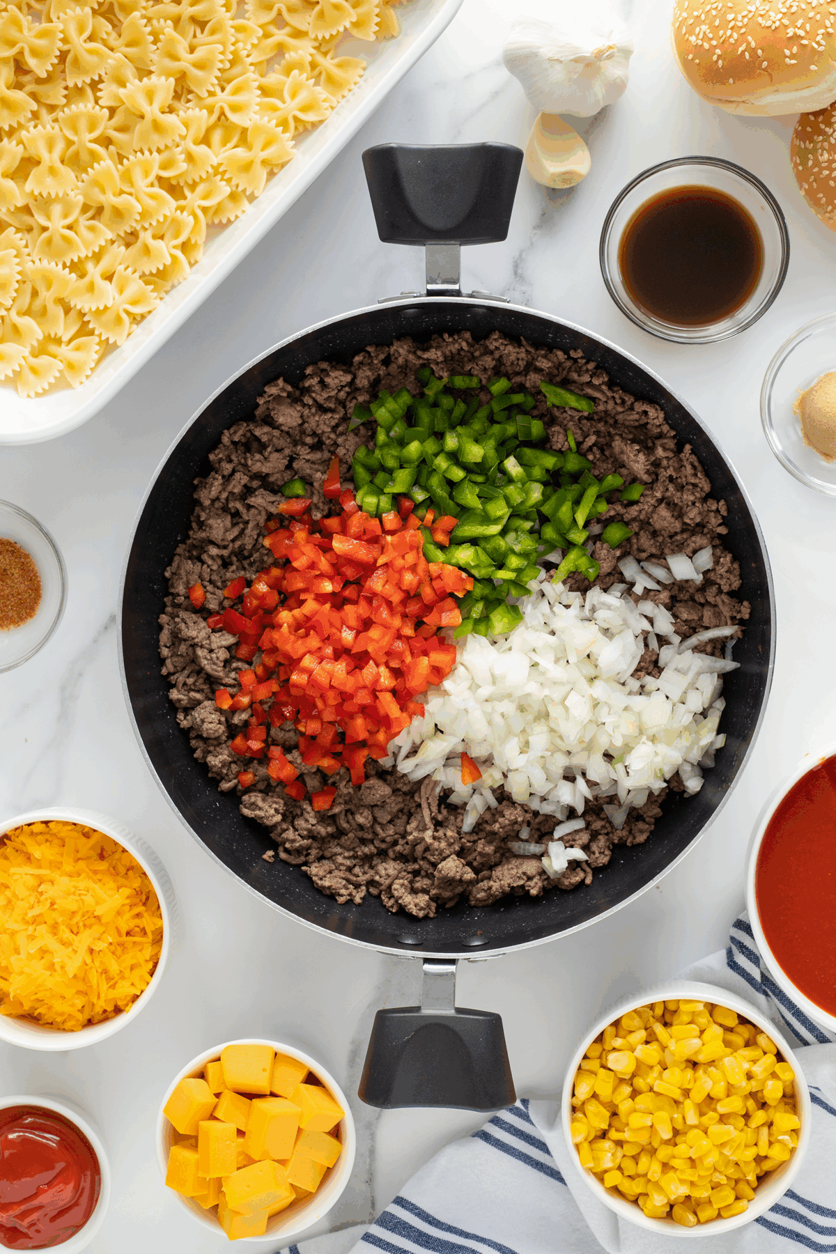 A skillet with cooked ground beef, topped with diced red bell pepper, green bell pepper, and onion, surrounded by bowls of pasta, cheese, corn, seasonings, and sauce on a white countertop.