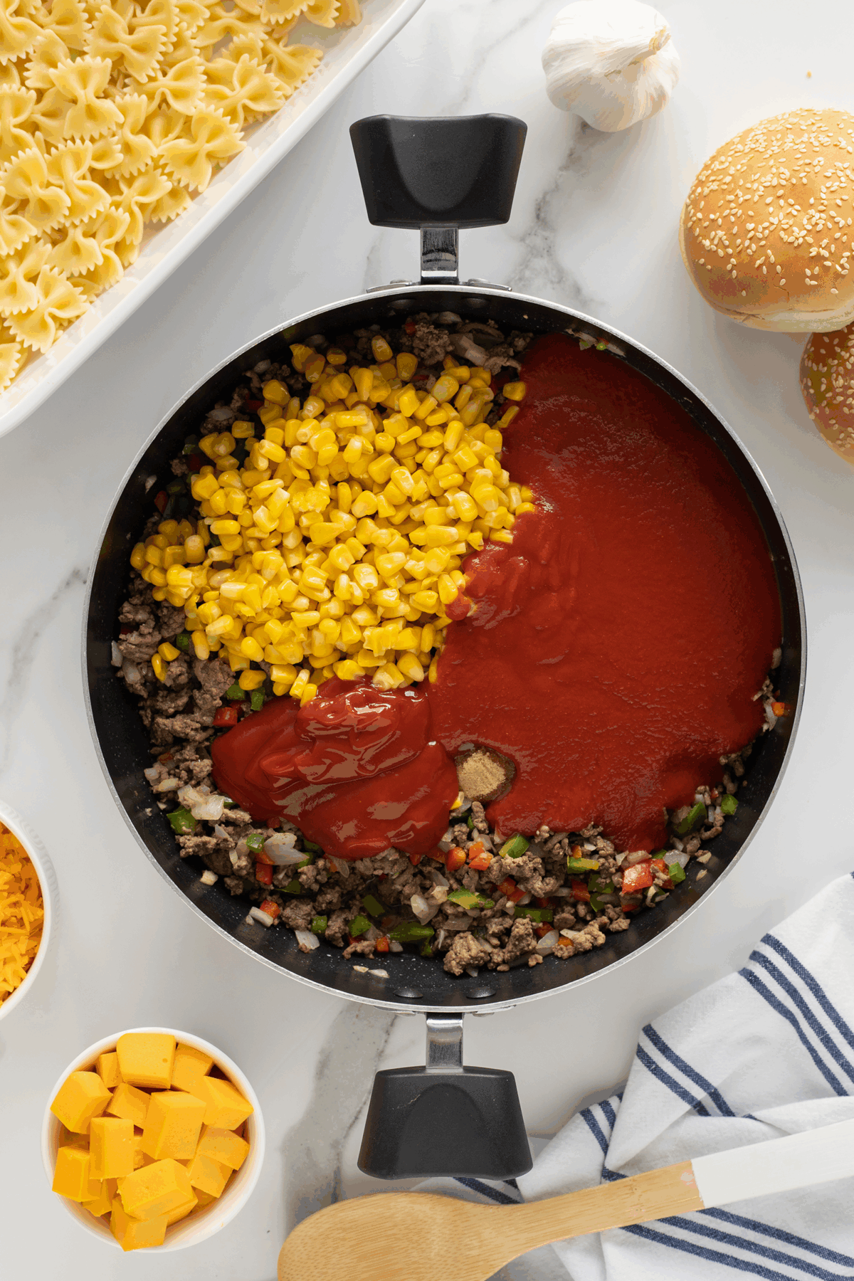 A skillet contains cooked ground beef with chopped vegetables, topped with corn, tomato sauce, and ketchup. Surrounding the skillet are uncooked farfalle pasta, cheese cubes, shredded cheese, hamburger buns, garlic, and a kitchen towel.