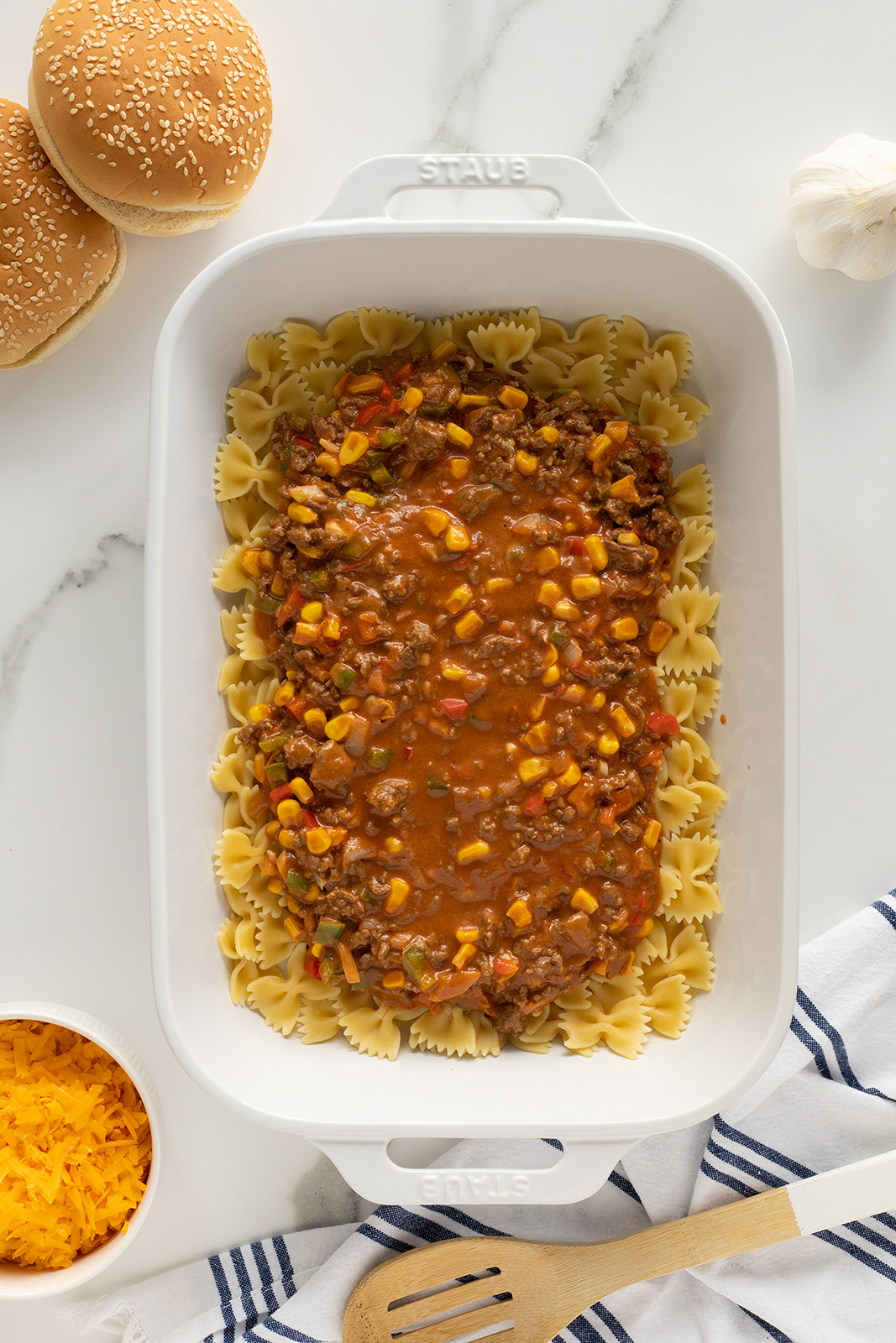 A white baking dish with a layer of bowtie pasta topped with ground beef, corn, and tomato sauce mixture. Surrounding the dish are burger buns, shredded cheese, a garlic bulb, and a wooden spoon on a striped towel.