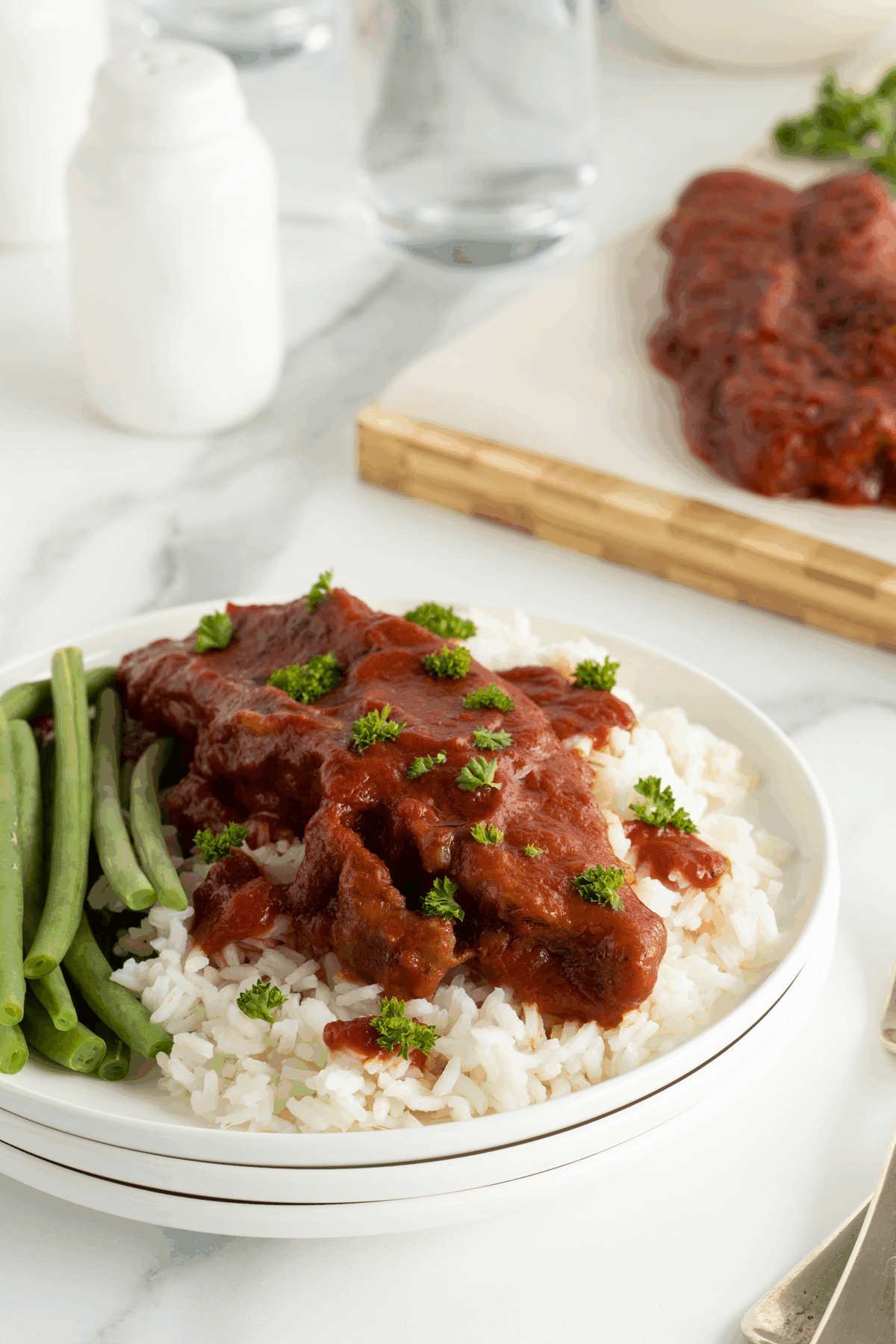 A plate of white rice topped with beef in tomato sauce, garnished with parsley, served with green beans. In the background, more saucy beef is on a cutting board.