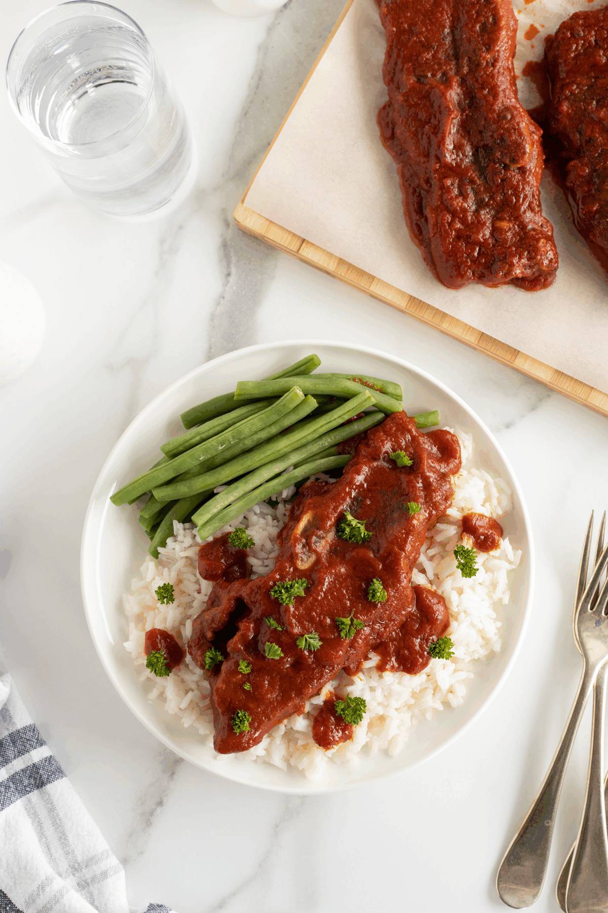 A plate of white rice topped with a slice of meat in tomato sauce, garnished with parsley, served alongside green beans. A glass of water, utensils, and extra sauced meat are nearby on a marble surface.