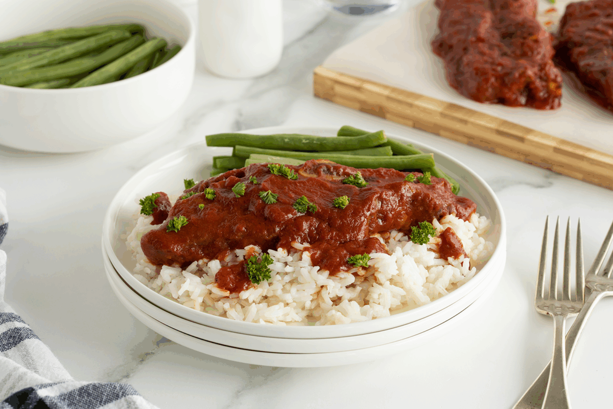 A plate of white rice topped with a saucy meat dish, garnished with parsley, and served with green beans. A bowl of green beans and extra meat with sauce are in the background on a marble surface.