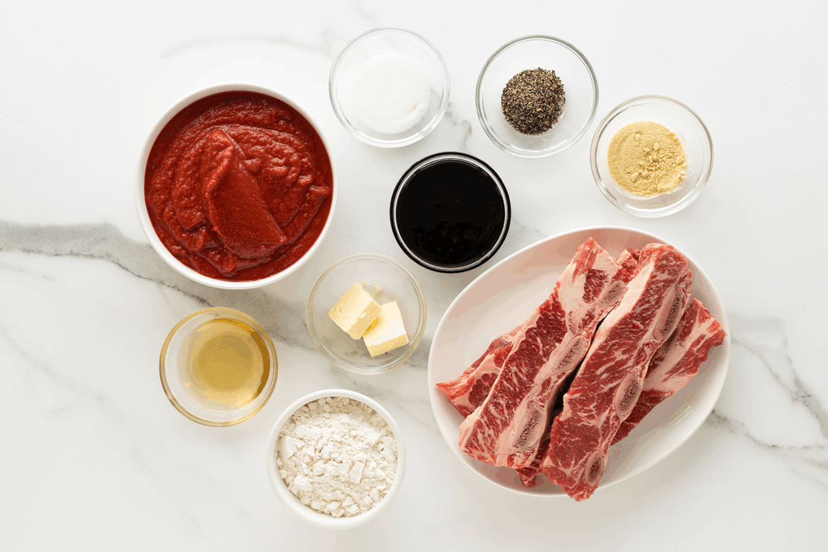 Overhead view of ingredients on a white surface: tomato sauce, white sugar, black pepper, ground mustard, brown beef short ribs, flour, butter, apple cider vinegar, and a dark liquid (likely Worcestershire sauce) in small bowls.