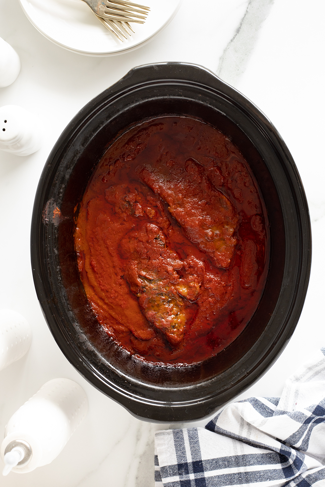 A black slow cooker filled with beef covered in tomato sauce sits on a white surface, with a blue and white plaid towel, white plates, forks, and salt and pepper shakers nearby.