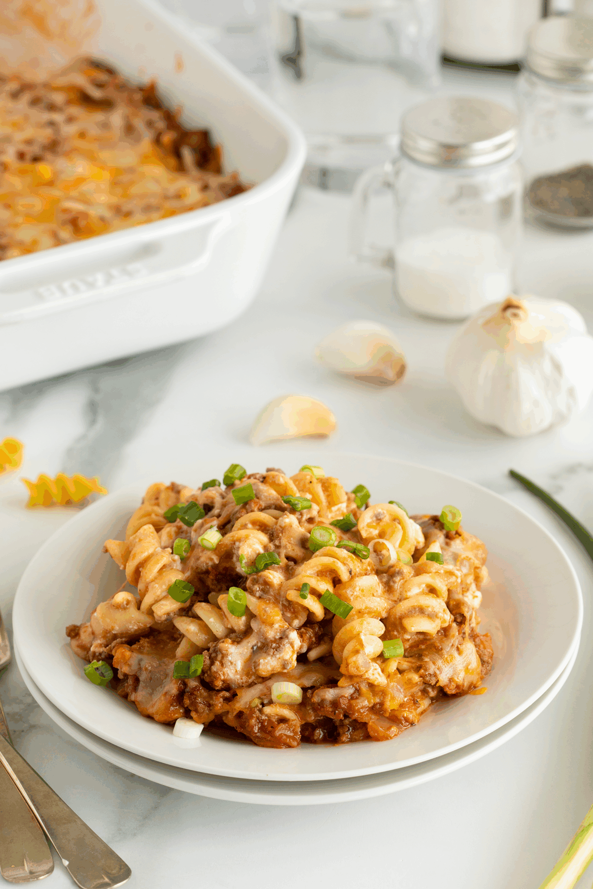 A serving of baked pasta with ground meat, melted cheese, and chopped green onions sits on a white plate, with garlic cloves, a casserole dish, and seasonings in the background.