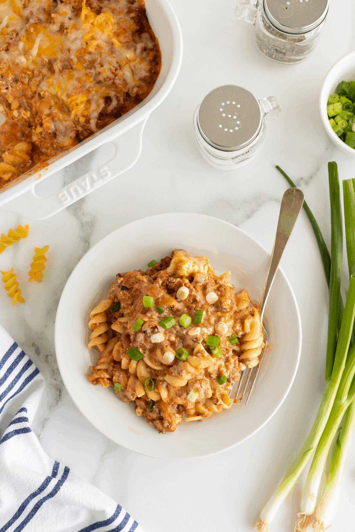 A plate of baked pasta with ground meat, sauce, and melted cheese, garnished with chopped green onions, sits next to a casserole dish of the same pasta. A fork, green onions, and seasoning shakers are nearby on a white surface.