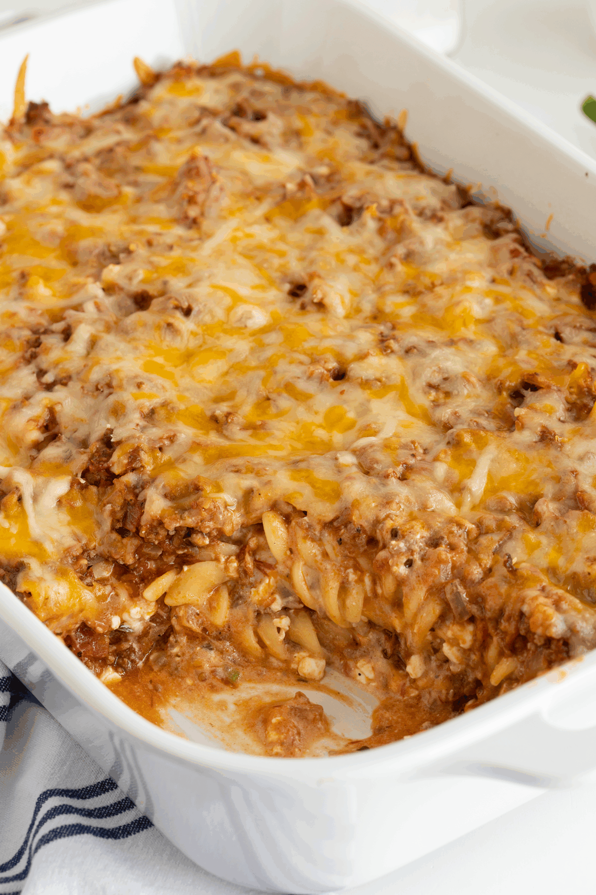 A baked casserole dish filled with cheesy, browned beef pasta bake, with a section scooped out, revealing layers of pasta, meat sauce, and melted cheese on top.