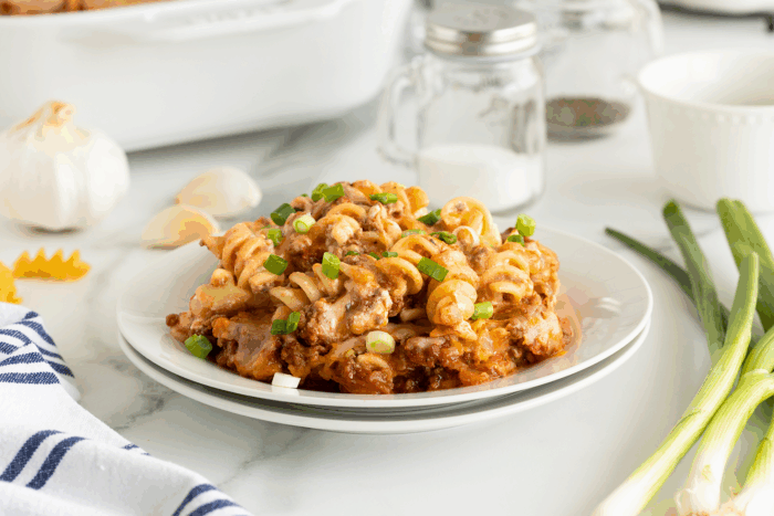 A plate of baked rotini pasta with ground beef, tomato sauce, melted cheese, and chopped green onions, set on a white table with garlic, green onions, and condiments in the background.