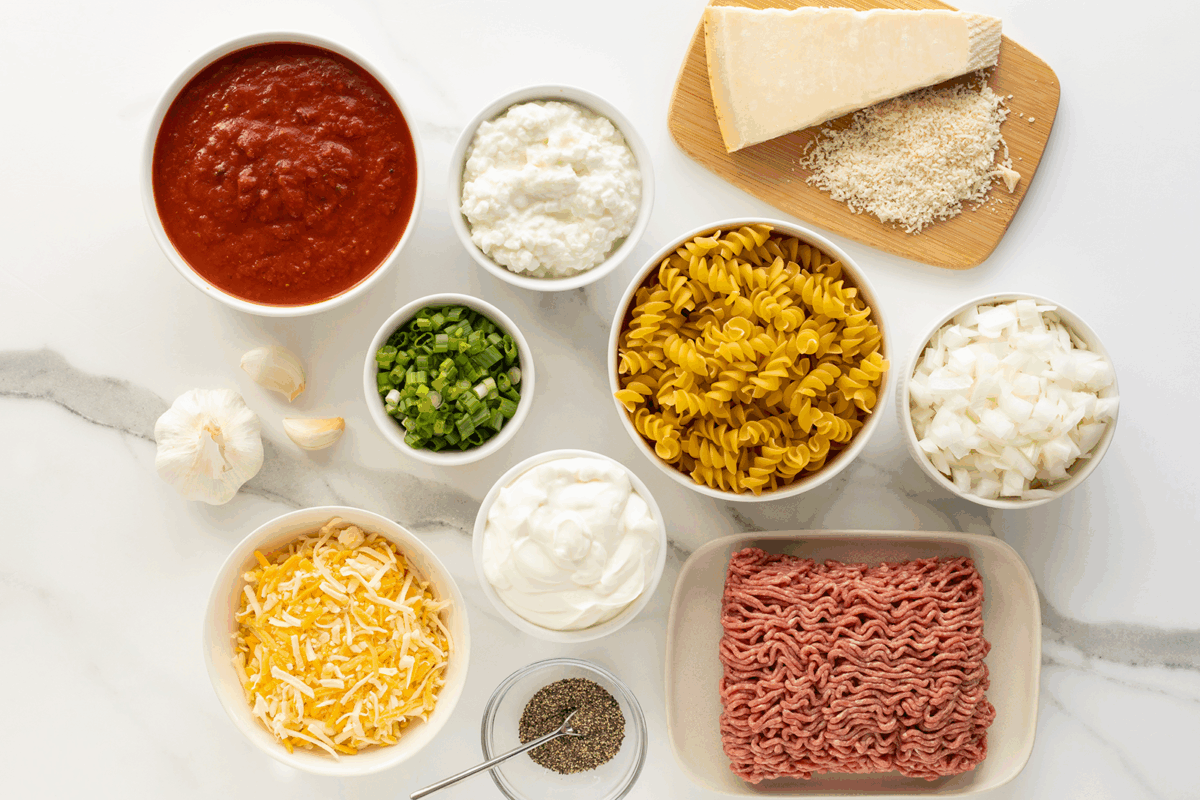 Bowls of ingredients for a pasta dish on a white countertop, including rotini, ground beef, chopped onions, green onions, shredded cheese, cottage cheese, marinara sauce, sour cream, garlic, pepper, and grated Parmesan.