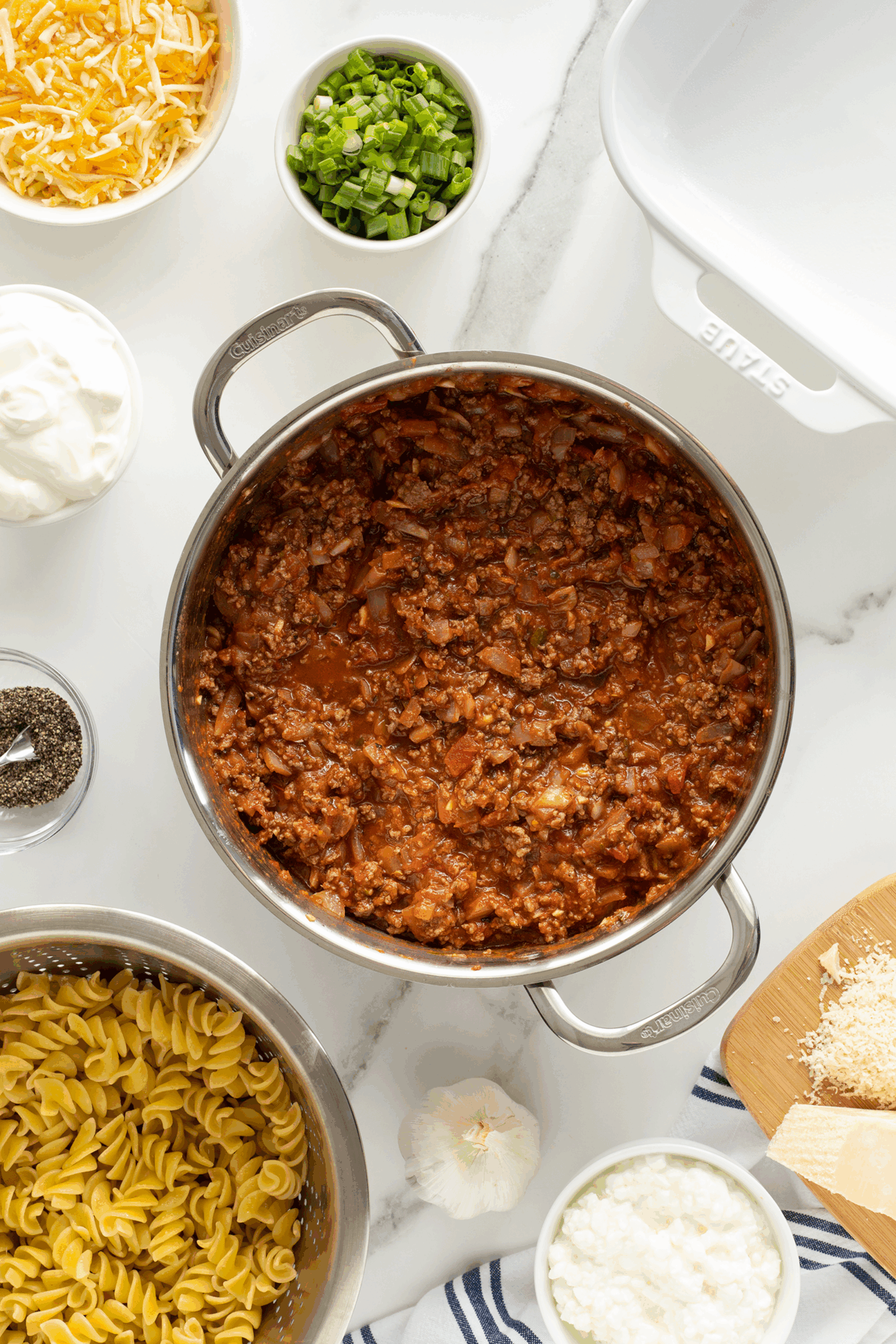 A pot of cooked meat sauce is surrounded by bowls of shredded cheese, sour cream, chopped green onions, cooked noodles, cottage cheese, black pepper, and a garlic bulb on a marble countertop. An empty baking dish is at the top right.