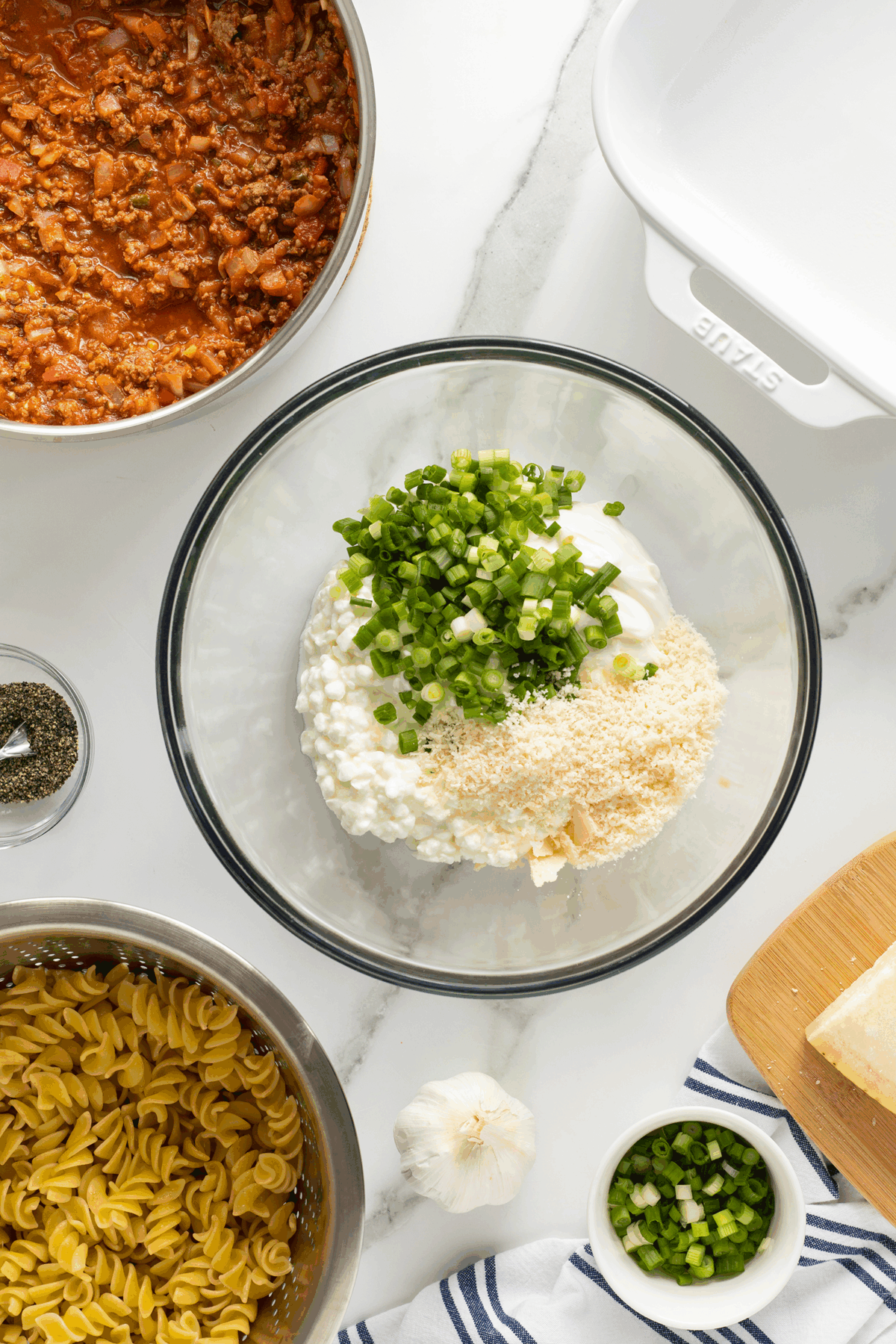 A glass bowl with cottage cheese, sour cream, breadcrumbs, and chopped green onions sits on a marble counter next to cooked pasta, a skillet of meat sauce, grated cheese, seasonings, and a baking dish.