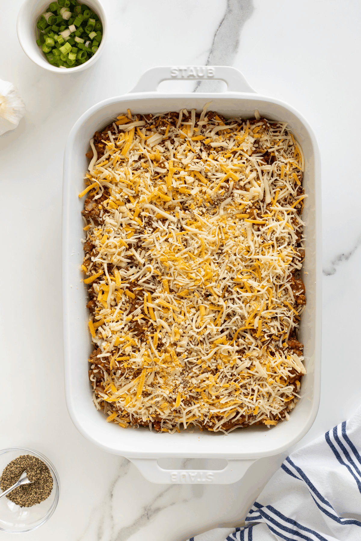 A white baking dish filled with an uncooked casserole topped with shredded cheese sits on a marble countertop, next to a bowl of sliced green onions, a bowl of ground pepper, and a striped kitchen towel.