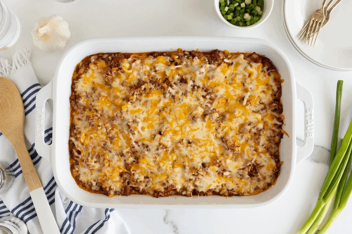 A baked casserole topped with melted cheese in a white dish, surrounded by a wooden spoon, a towel, green onions, garlic, plates, forks, and a bowl of sliced green onions on a white surface.