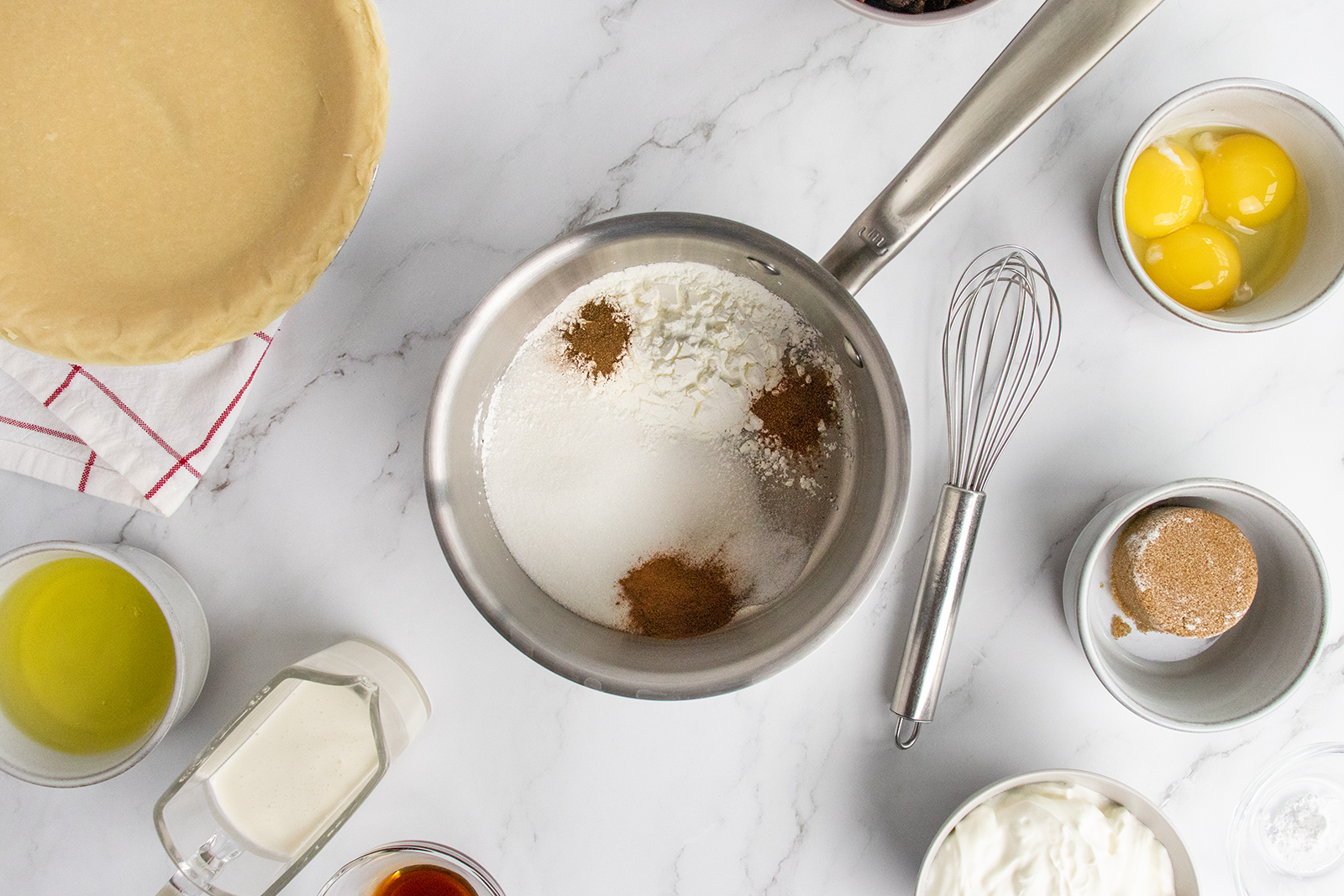 A marble countertop displays a pie crust, a saucepan with dry ingredients, a whisk, eggs, brown sugar, butter, cream, vanilla, and sour cream, all arranged for baking preparation.