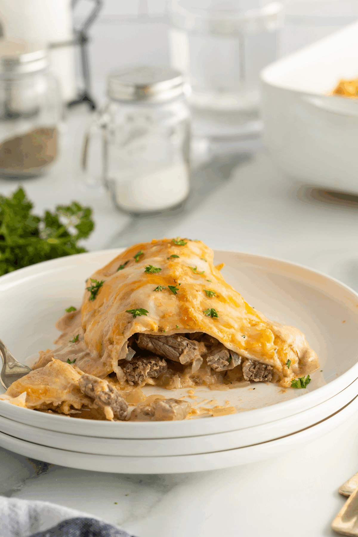 A close-up of a cheesy beef enchilada topped with melted cheese and chopped parsley on a white plate, with a fork and garnishes in the background.