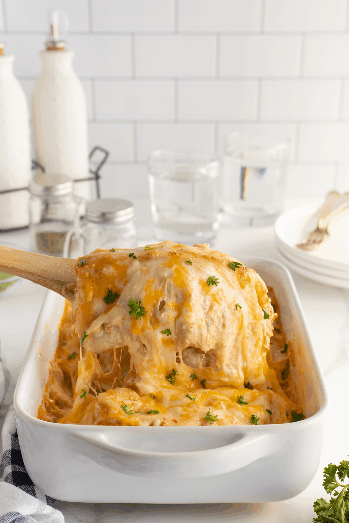A cheesy baked casserole is being served with a wooden spatula from a white baking dish, topped with melted cheese and fresh herbs. Two glasses of water, plates, and utensils are in the background.