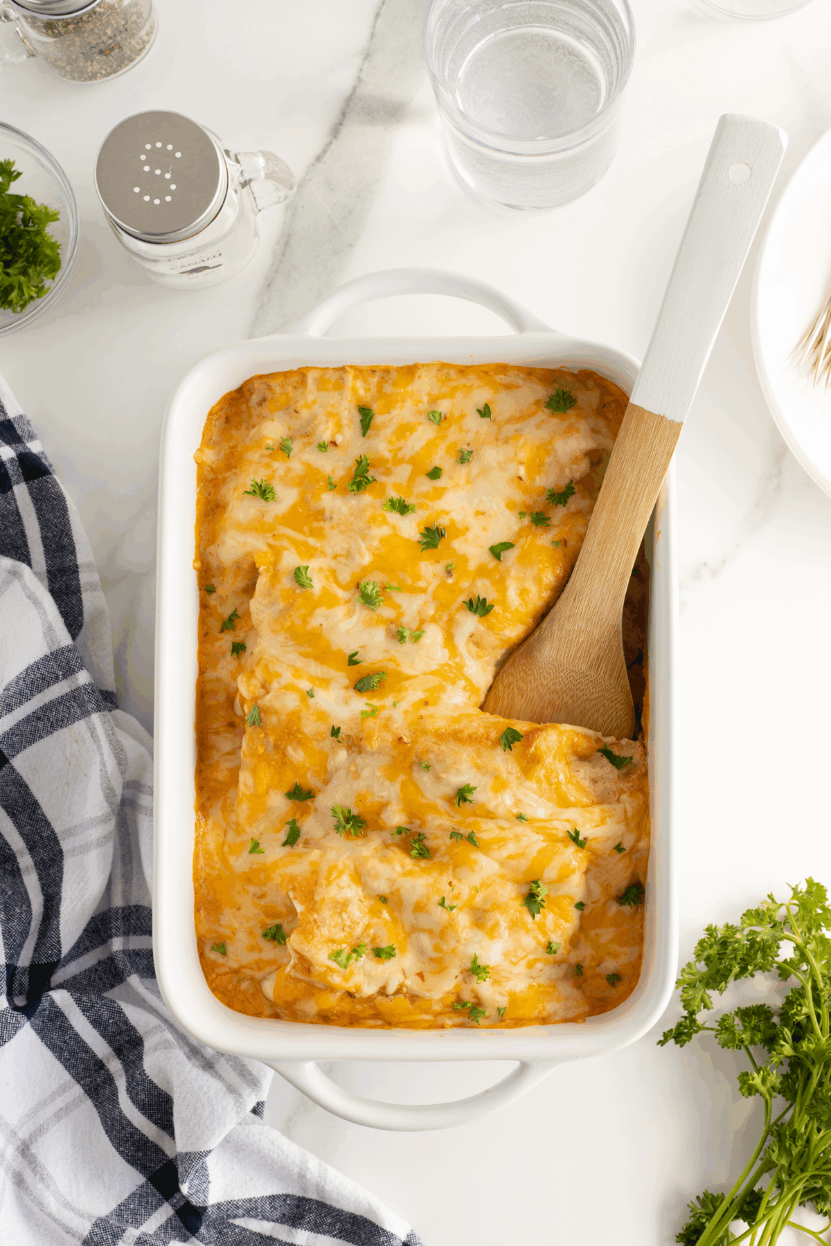 A white baking dish filled with cheesy baked casserole topped with chopped parsley. A wooden spoon rests in the dish. Nearby are a striped towel, fresh parsley, a glass of water, and seasonings on a white marble surface.