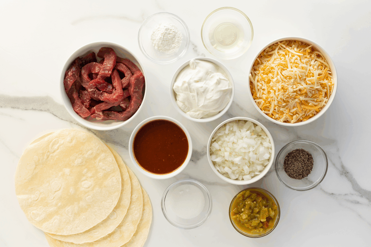 A top-down view of ingredients for beef enchiladas, including sliced beef, flour tortillas, shredded cheese, sour cream, diced onions, green chilies, enchilada sauce, black pepper, flour, and oil in separate bowls.