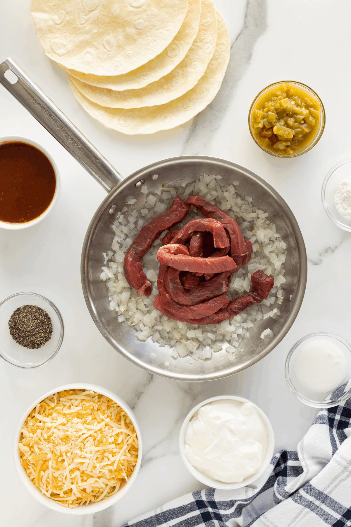 A skillet with sliced raw beef and chopped onions is surrounded by tortillas, shredded cheese, sour cream, black pepper, green chiles, flour, and enchilada sauce on a white marble countertop.