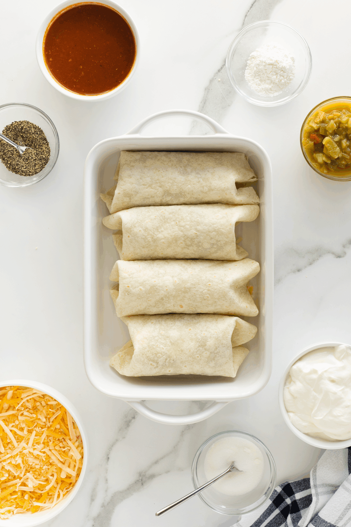 A white baking dish with four wrapped burritos is surrounded by bowls of red sauce, shredded cheese, black pepper, flour, green chilies, sour cream, and a whisk on a marble surface.