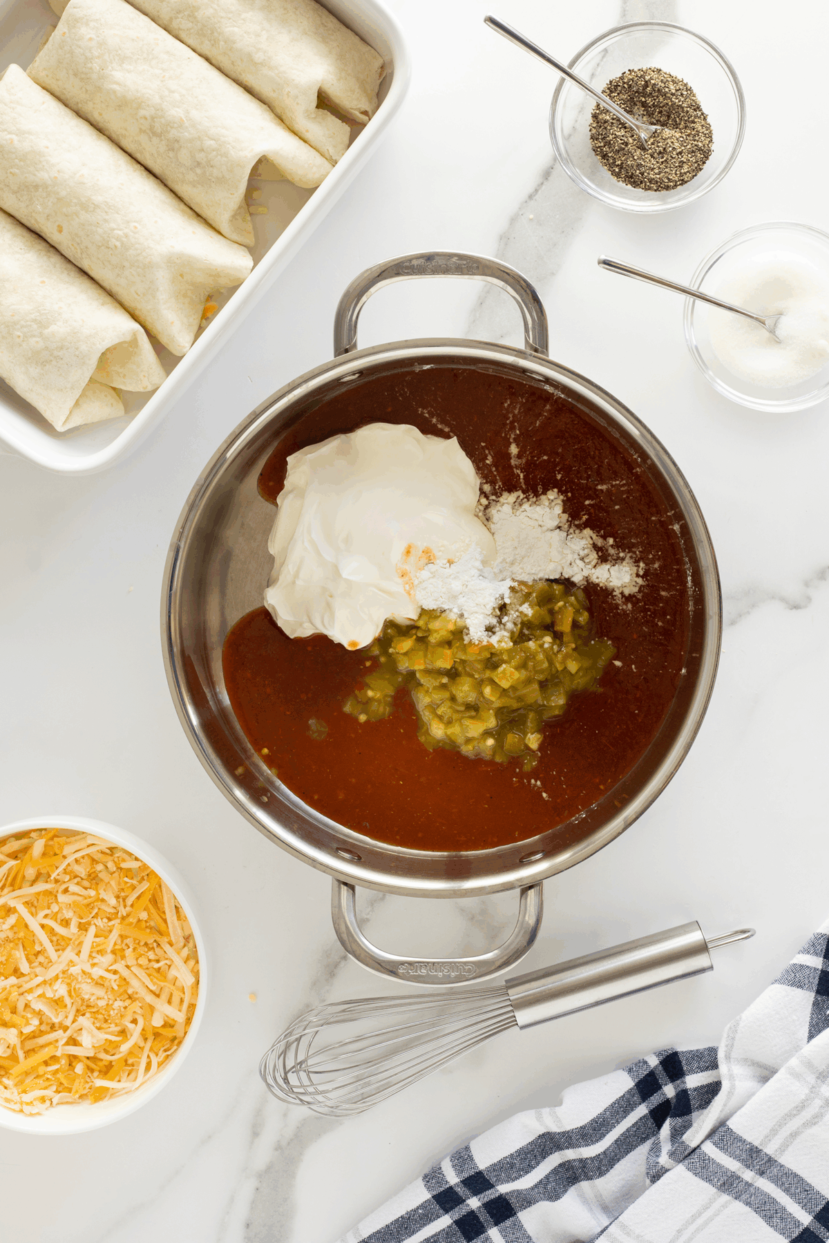 A saucepan with enchilada sauce, sour cream, flour, and green chiles, surrounded by a whisk, shredded cheese, rolled tortillas in a baking dish, and small bowls of salt and pepper on a white countertop.