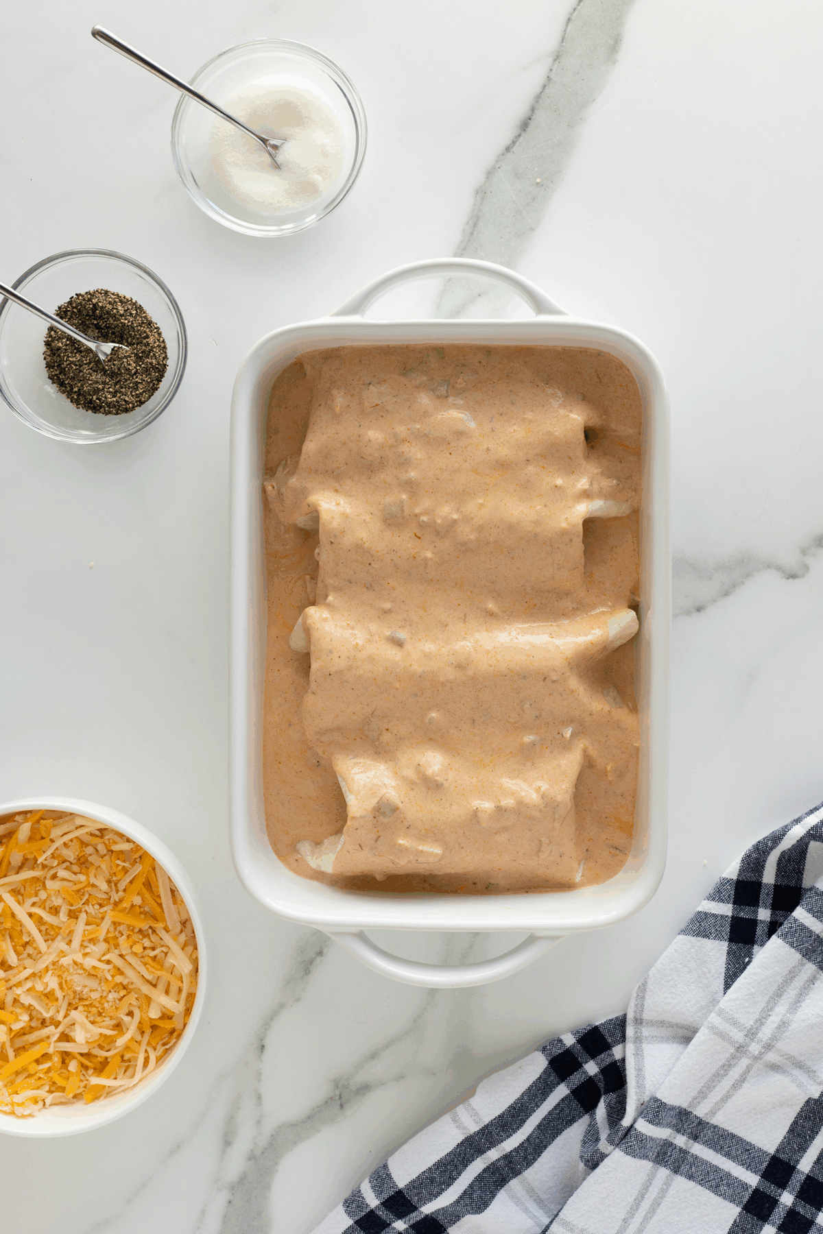 A white baking dish with enchiladas covered in a creamy, orange sauce sits on a marble countertop. Surrounding the dish are bowls of shredded cheese, black pepper, salt, and a blue plaid towel.