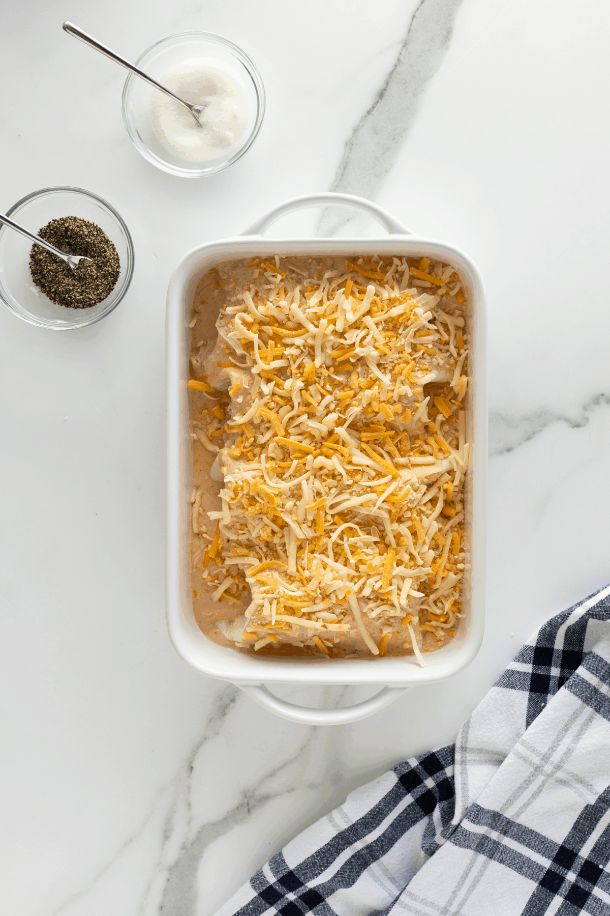 A white baking dish filled with shredded cheese over a casserole sits on a marble counter, next to small bowls of ground pepper and salt with spoons, and a folded black-and-white checked kitchen towel.