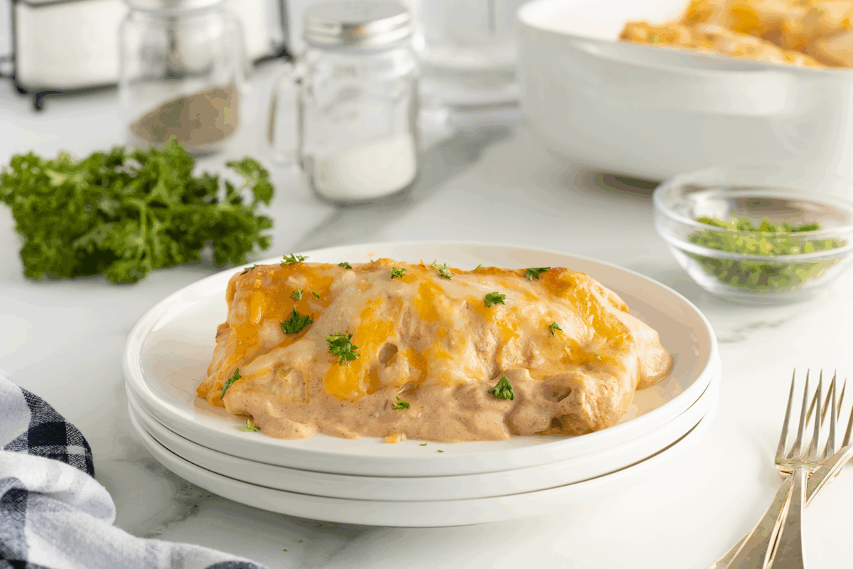 A cheesy enchilada topped with melted cheese and fresh parsley sits on a white plate. In the background, there are ingredients including a bunch of parsley, seasonings, and a dish of more enchiladas.