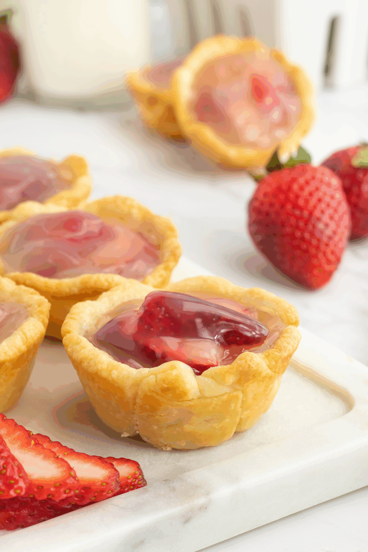 Mini tart shells filled with glossy strawberry topping are arranged on a white surface, with fresh whole and sliced strawberries nearby. More tarts and a glass of milk appear in the background.