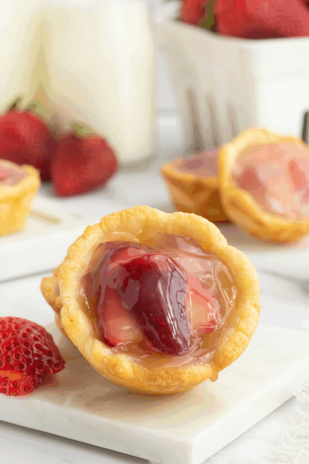 A close-up of a mini tart filled with glazed strawberries and custard, with whole strawberries and more tarts in the background on a white surface.