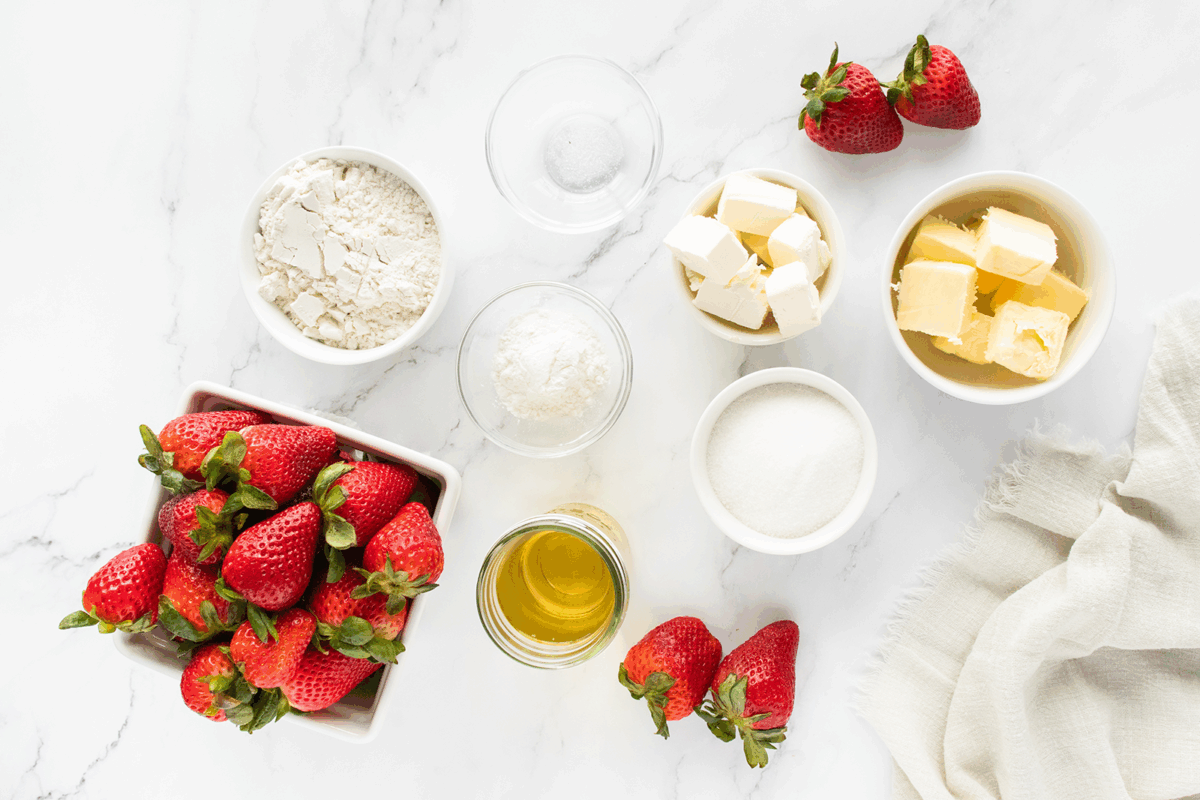Fresh strawberries, flour, powdered sugar, granulated sugar, cubed butter, cubed cream cheese, honey in a jar, and a pinch of salt arranged neatly on a white marble surface with a beige cloth nearby.