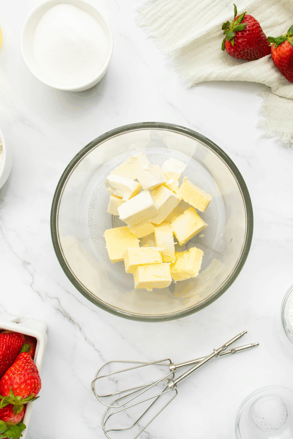 A glass bowl with cubed butter on a marble surface, surrounded by strawberries, a bowl of sugar, and metal beaters.