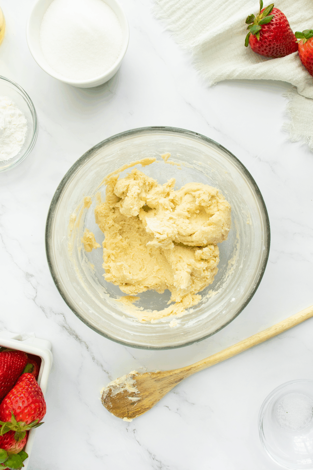 A glass bowl filled with cookie dough sits on a marble countertop, surrounded by a wooden spoon, fresh strawberries, a bowl of sugar, a bowl of flour, and a white cloth.