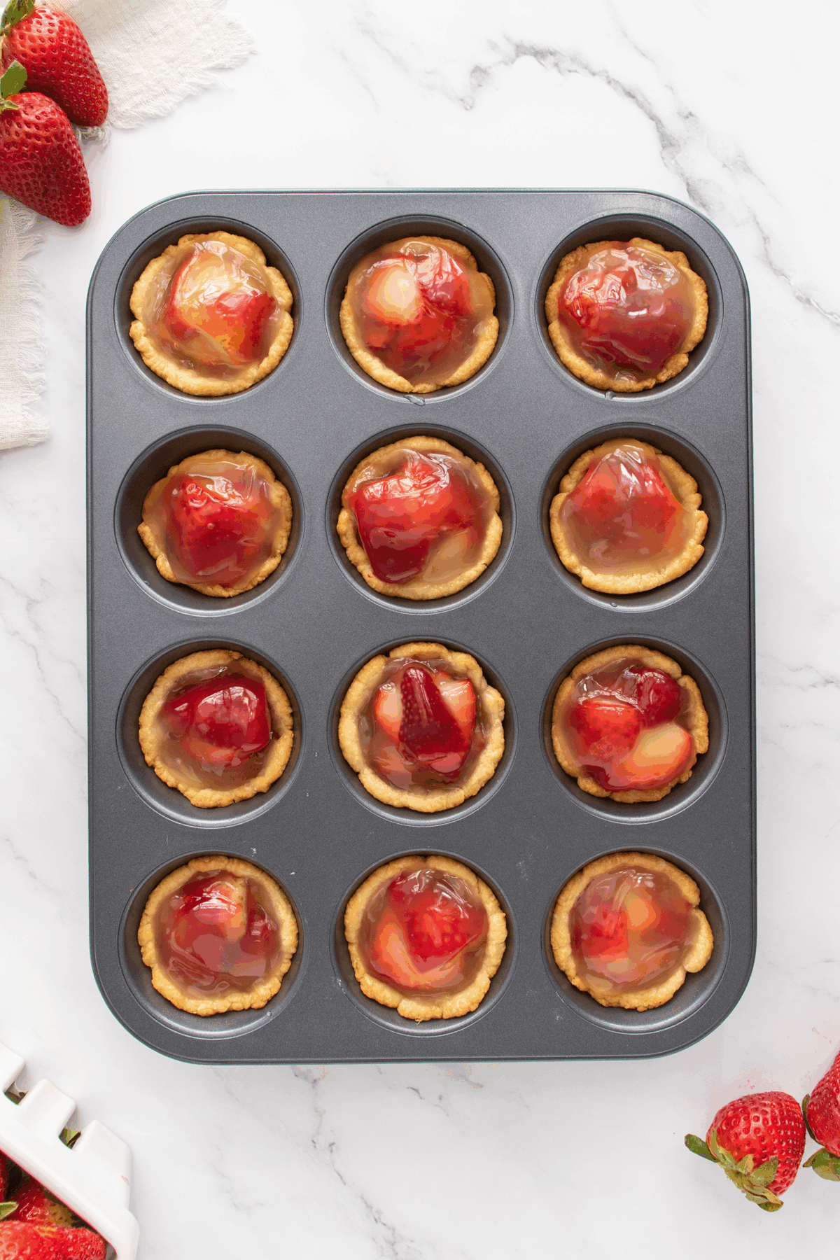 A muffin tin filled with twelve mini strawberry tarts, each with a glossy strawberry topping. Fresh strawberries are scattered on a white marble surface around the tin.