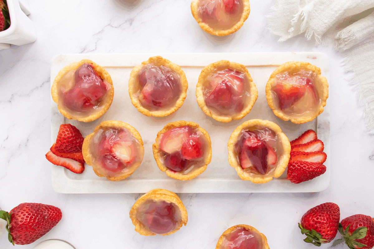 Nine mini tartlets filled with strawberries and a glossy glaze are arranged on a rectangular white tray, with fresh strawberry slices and whole strawberries placed around for decoration.